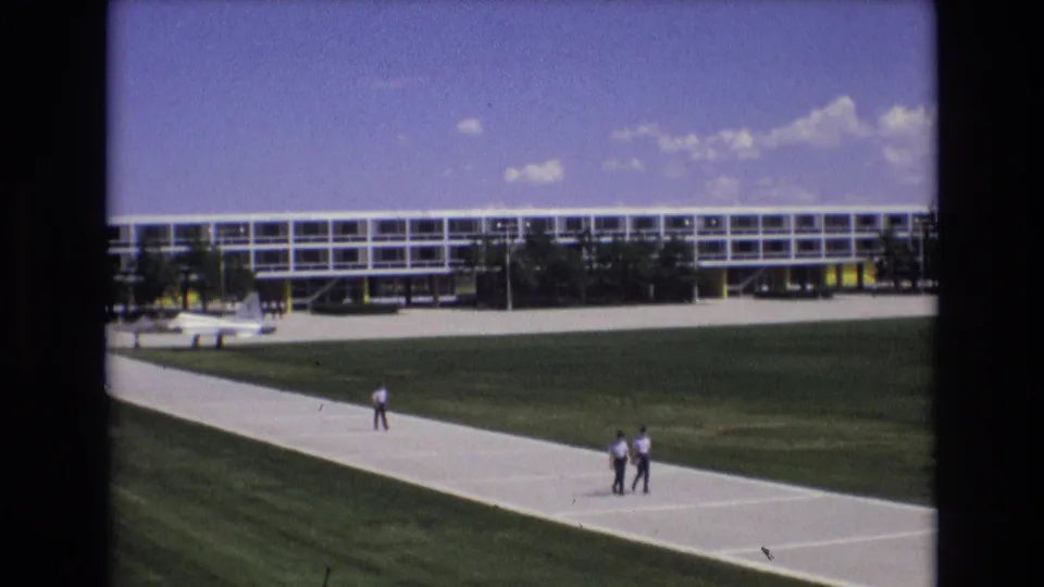 1973 Colorado Springs 8mm frame showing two men near the lower walkway and another distant person farther along the path, with a long modern building behind the lawn.