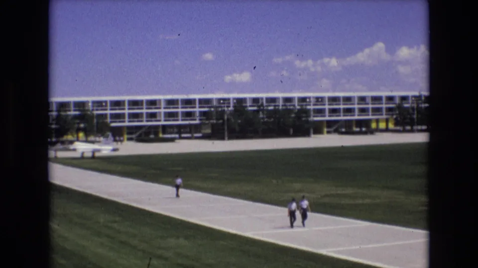 1973 Colorado Springs frame with two men walking along the right side of the paved walkway, a third distant figure behind them, and the long building spanning the background.