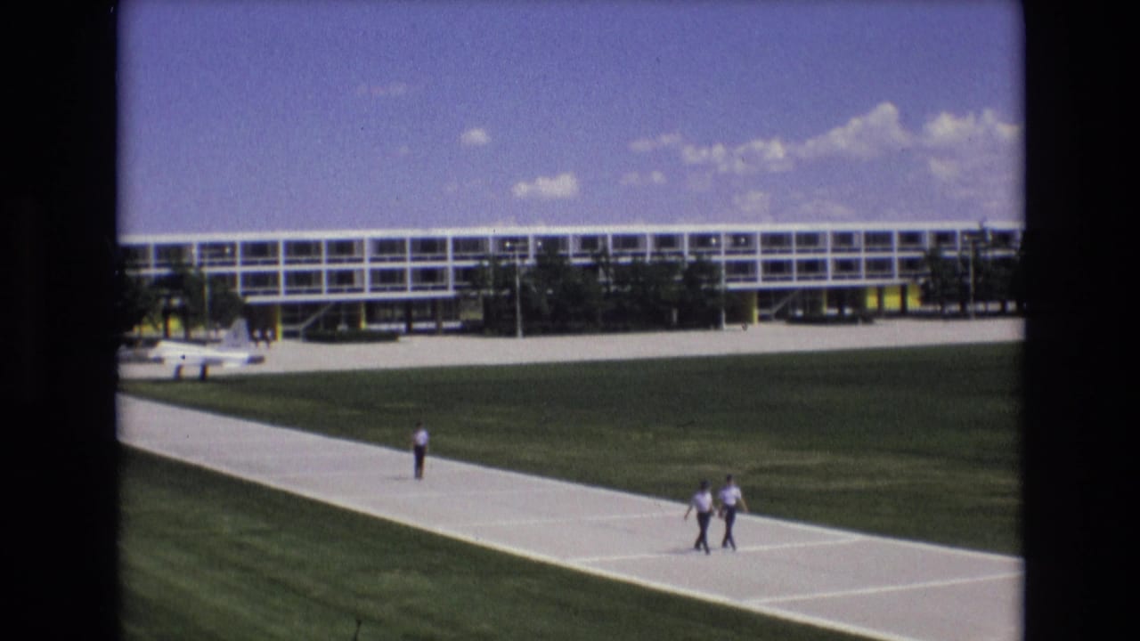 1973 Colorado Springs home-movie frame showing a long grid-window building, green lawns, broad concrete walkways, and several distant people walking outdoors.