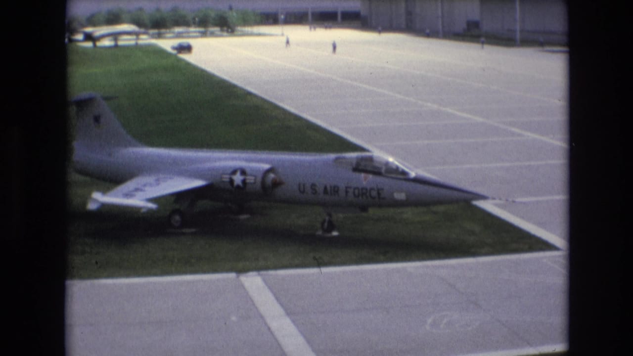 1973 Colorado Springs frame showing a U.S. Air Force-marked jet displayed on grass next to broad concrete pavement, with small distant figures and buildings in the background.