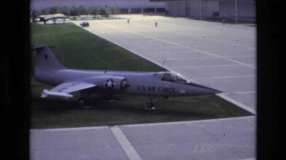 1973 Colorado Springs home-movie frame with a side view of a U.S. Air Force jet on display beside a wide paved area and green lawn.