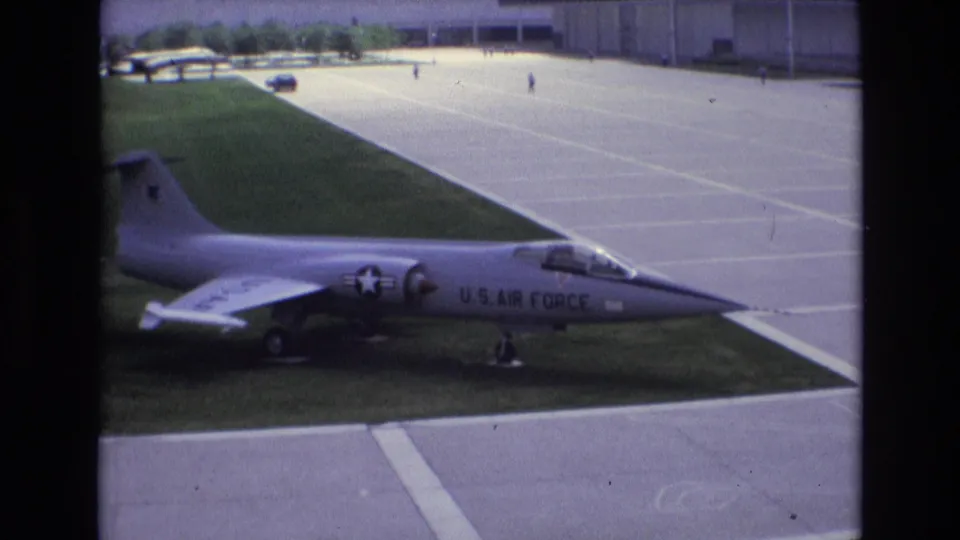 1973 Colorado Springs frame showing the displayed U.S. Air Force jet from the side, with grass, concrete pavement, distant figures, and hangar-like buildings behind it.
