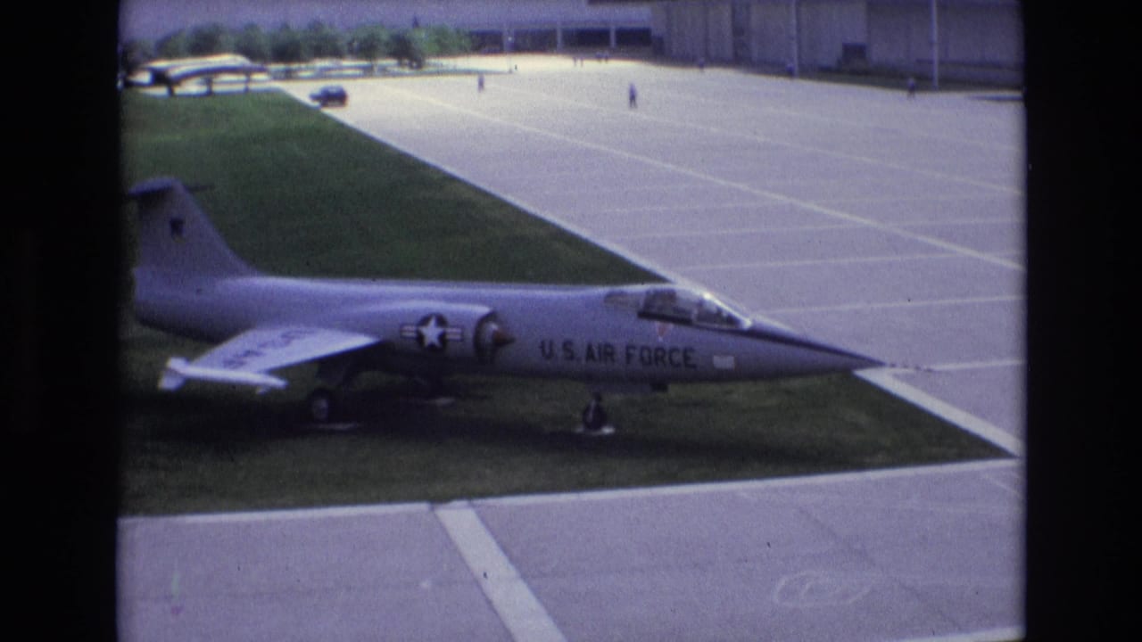 A silver U.S. Air Force-marked jet sits on a grass strip next to broad concrete pavement in metadata-supported 1973 Colorado Springs footage.