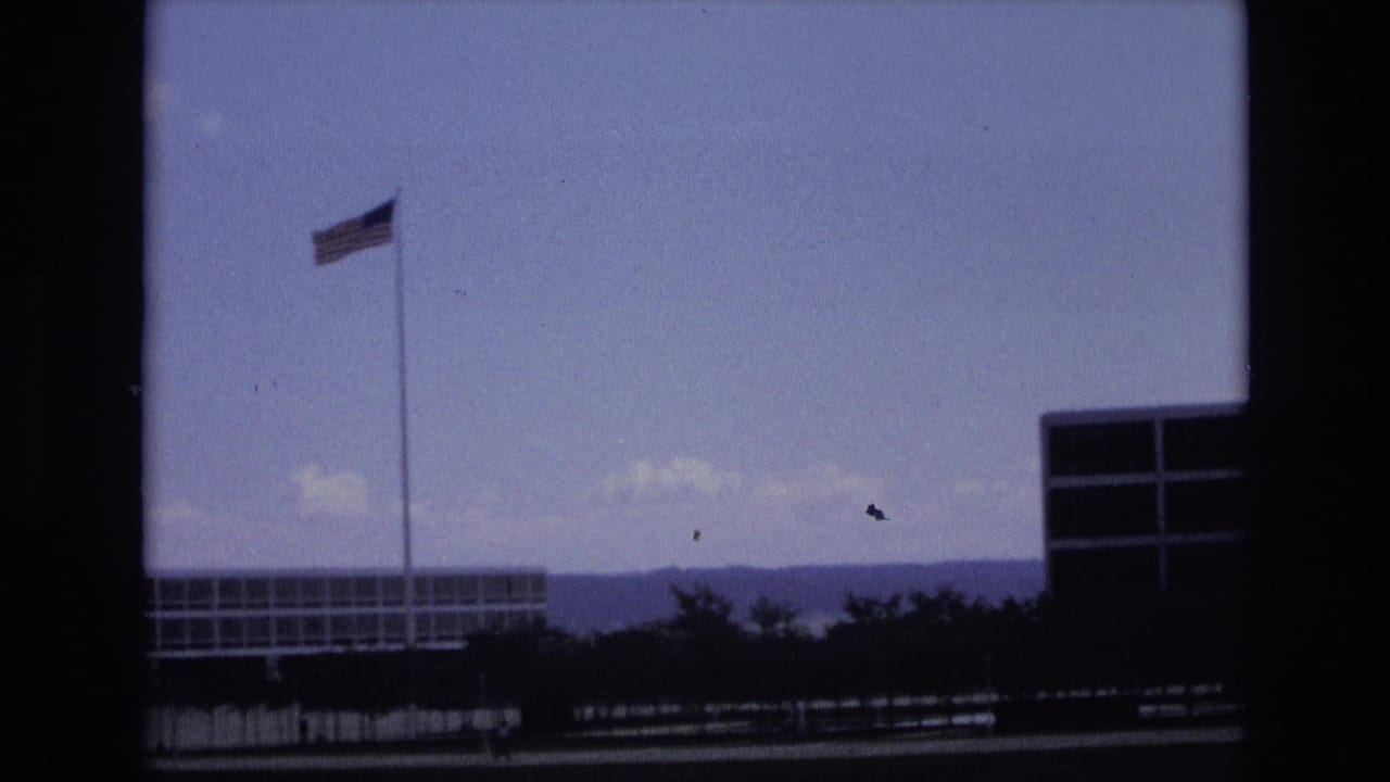 The camera frames a tall American flagpole, low base buildings, and distant hills in the metadata-supported Colorado Springs setting.