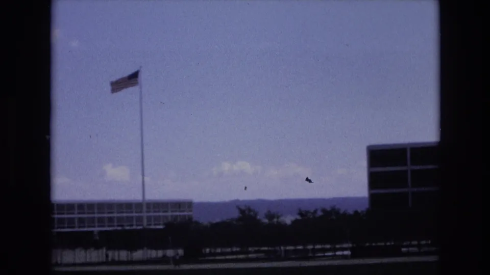 The camera frames a tall American flagpole, low base buildings, and distant hills in the metadata-supported Colorado Springs setting.