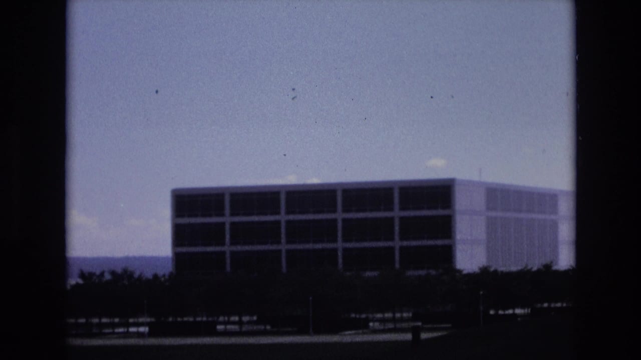 1973 Colorado Springs home-movie frame showing a large blocky modernist building with dark window bands, trees in the foreground, distant hills, and a clear blue sky.