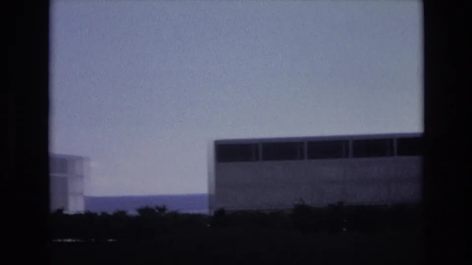 1973 Colorado Springs home-movie frame showing a partial concrete building at left, a larger cropped modernist structure at right, low trees, distant hills, and pale blue sky.