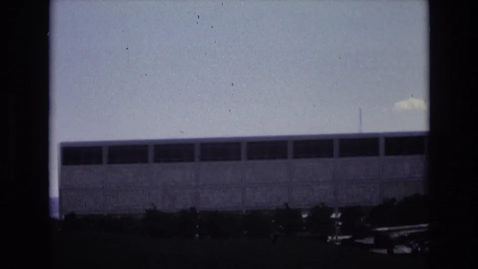 1973 Colorado Springs frame showing a long concrete-and-glass building, dark window banding, low trees, a shadowed roadside or parking area, and a small glimpse of vehicles at the edge.