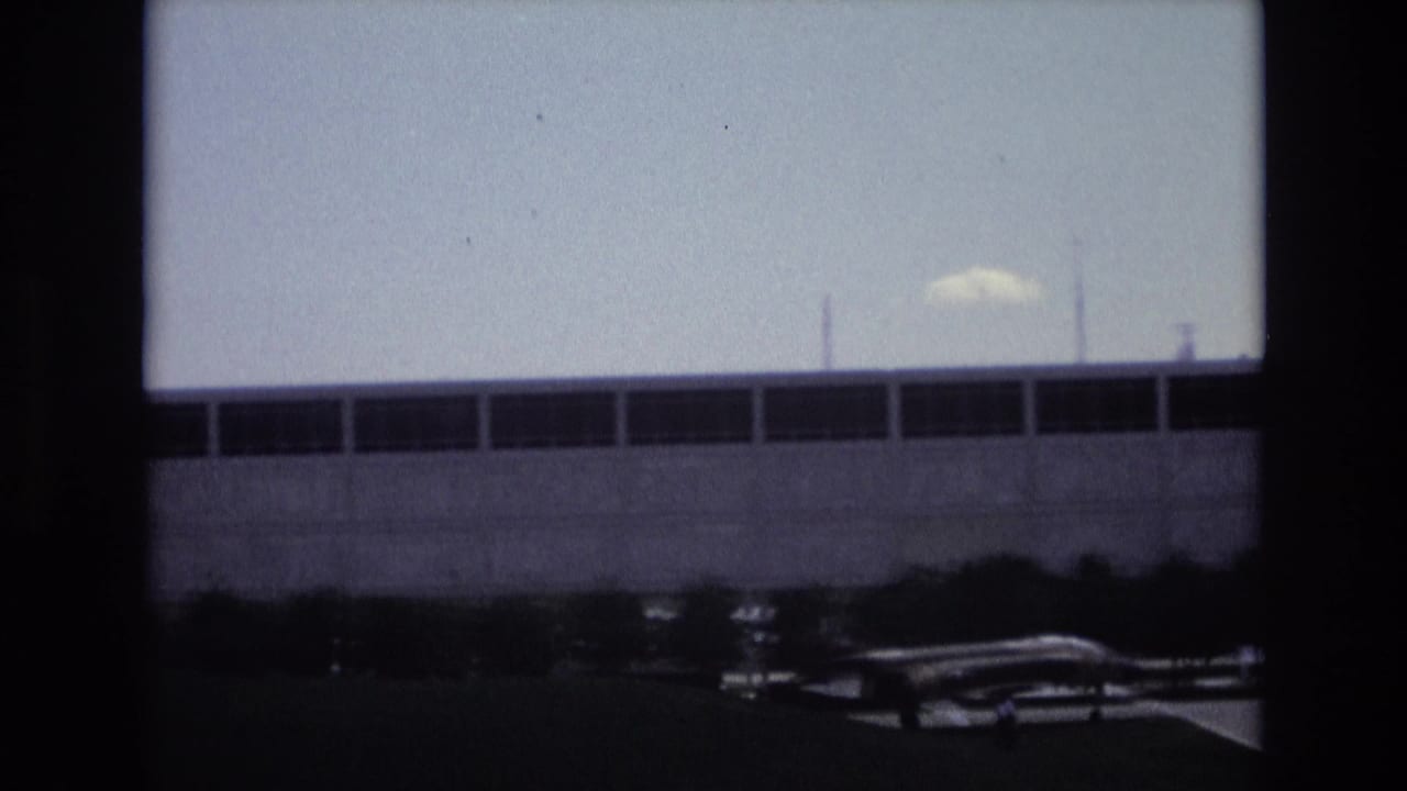 1973 Colorado Springs home-movie frame showing a large modern institutional building beyond a small aircraft, with dark film borders and a blue-gray 8mm look.