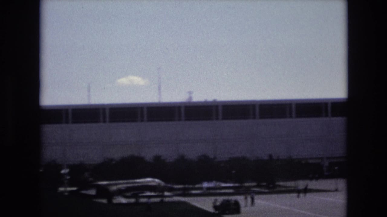 Wide exterior view of a large building, small aircraft, and distant people on pavement.