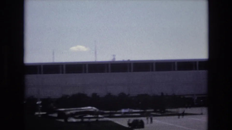 1973 Colorado Springs frame with a large windowed building, a small aircraft in the foreground, and distant people crossing the paved area.