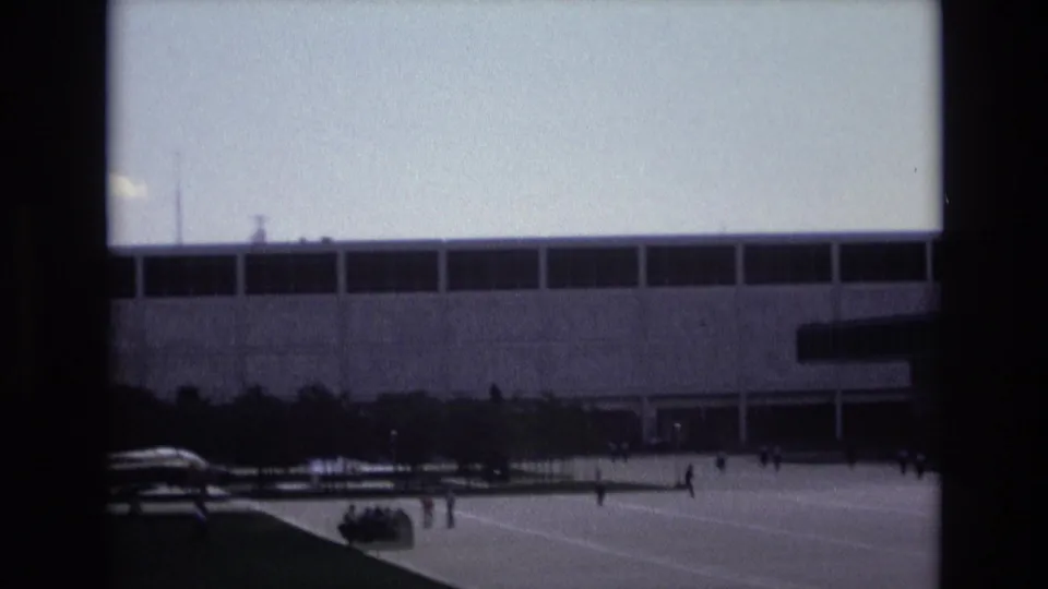 1973 Colorado Springs home-movie view of a broad modern building and small figures walking or running across the open paved area in front.