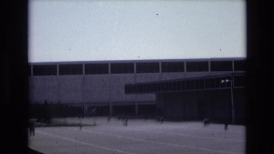 1973 Colorado Springs frame showing a large modern building and adjacent structure across an open paved plaza with small distant figures in motion.