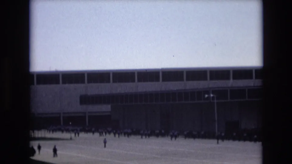 Colorado Springs 1973 home-movie still with distant people along a building frontage, a tall light pole, and a broad empty paved foreground.