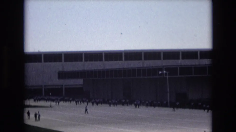 Final 1973 Colorado Springs frame showing scattered walkers and a distant organized group outside a large modern building, viewed through soft 8mm film texture.