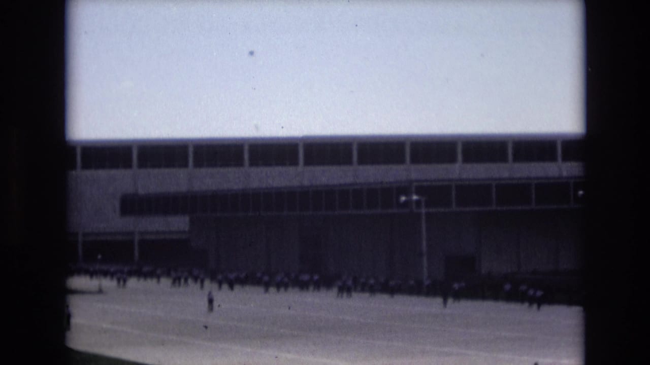 Distant 1973 Colorado Springs home-movie view of personnel on a broad paved drill or airfield area before a large grid-windowed institutional building.