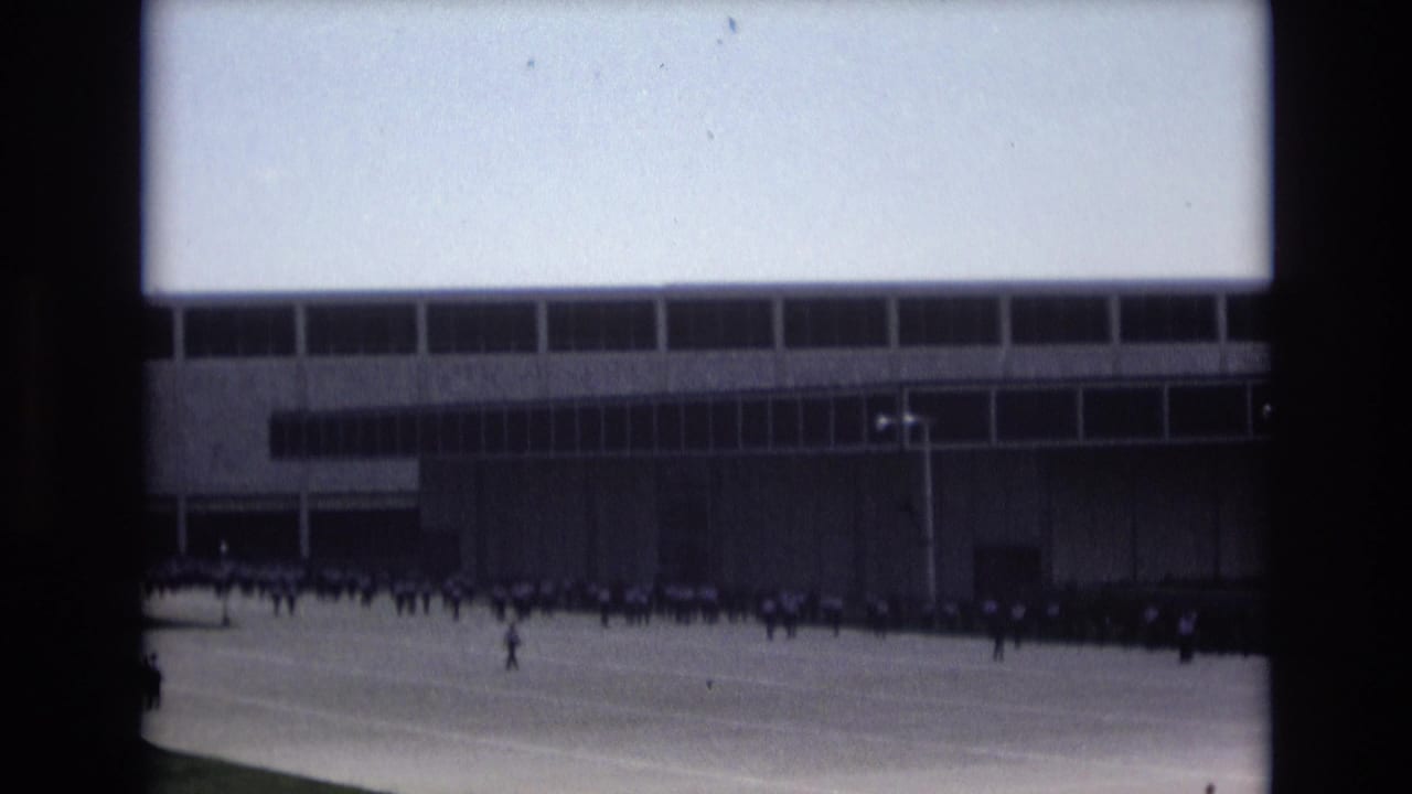 Small figures stand and move across pavement beside a large windowed building.