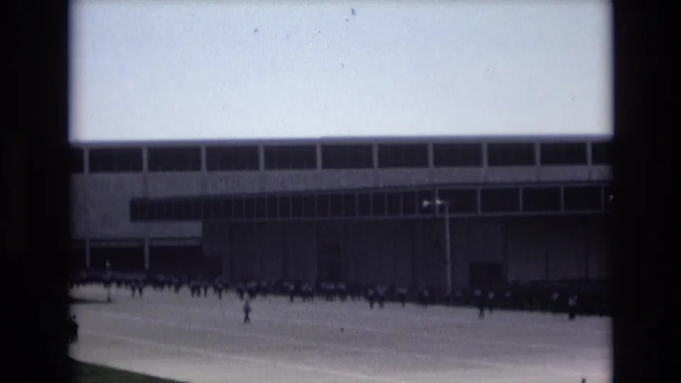 Distant home-movie frame of personnel moving across pavement near a large modernist building in the supplied Colorado Springs 1973 setting.