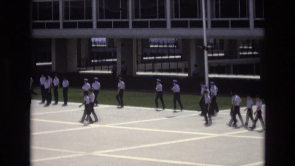 Closer view of uniformed personnel in white shirts and dark trousers walking across marked pavement outside a windowed institutional building in Colorado Springs, 1973.