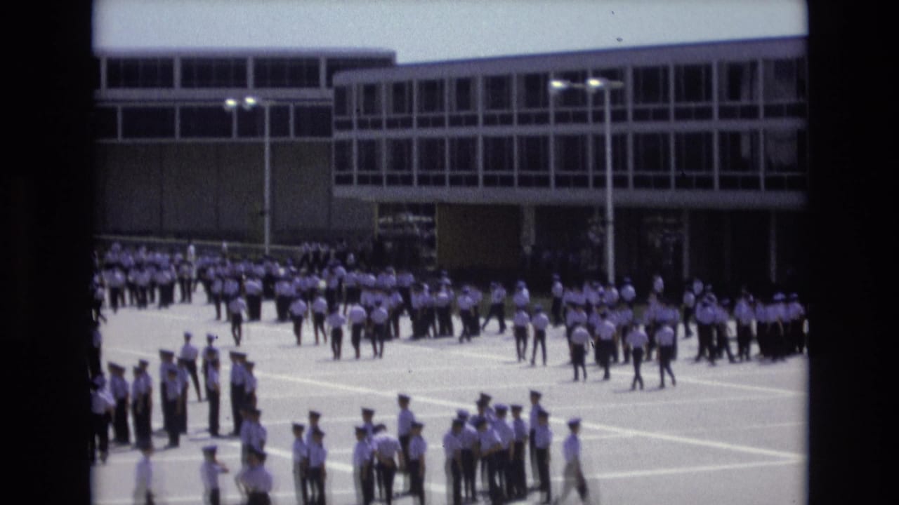 In 1973 Colorado Springs footage, uniformed cadets in white shirts and dark trousers assemble across a paved courtyard outside a large modern institutional building.