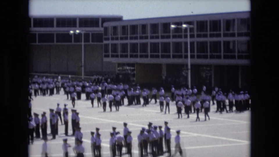 In 1973 Colorado Springs footage, uniformed cadets in white shirts and dark trousers assemble across a paved courtyard outside a large modern institutional building.