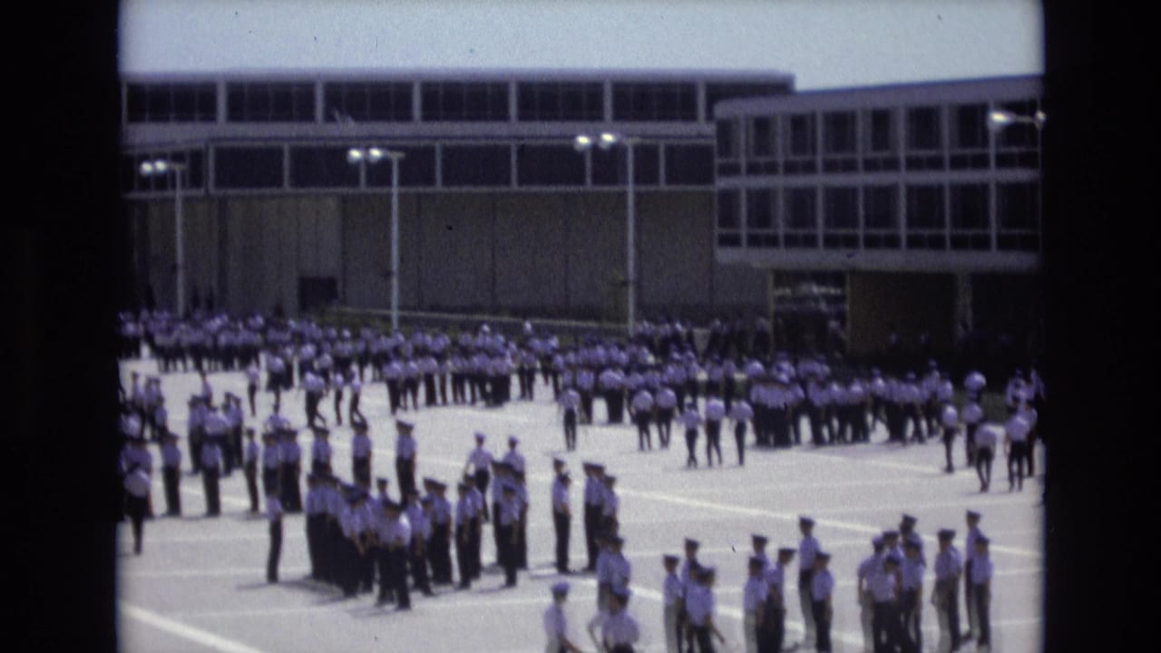 Uniformed cadets stand in ordered rows and small clusters on a bright paved courtyard in Colorado Springs, with a modern institutional building behind them.
