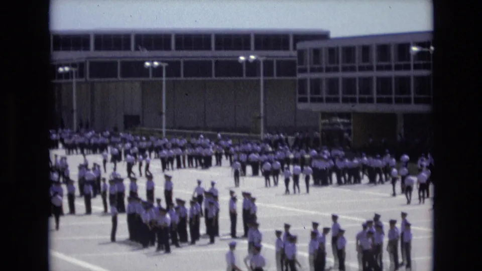 Uniformed cadets stand in ordered rows and small clusters on a bright paved courtyard in Colorado Springs, with a modern institutional building behind them.