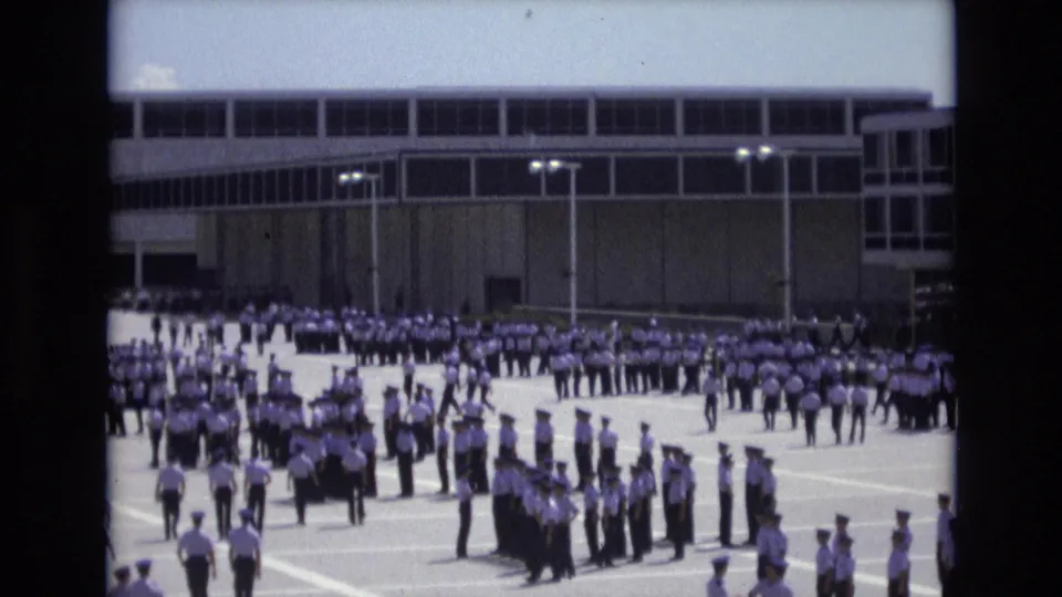 Multiple lines of cadets in white shirts and caps assemble in a paved courtyard, with other uniformed people walking into position beneath the large building facade.