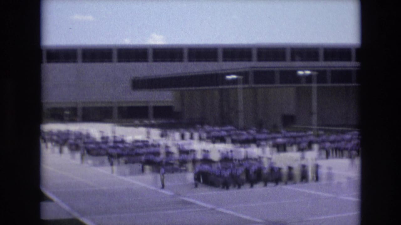 1973 Colorado Springs home-movie frame showing large groups of uniformed students standing in organized rows on a paved schoolyard before a windowed institutional building.