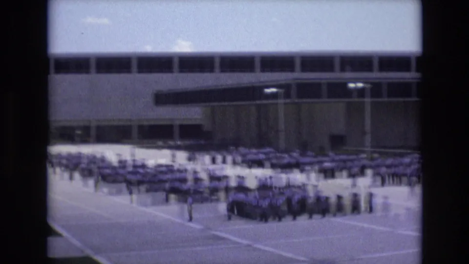 1973 Colorado Springs home-movie frame showing large groups of uniformed students standing in organized rows on a paved schoolyard before a windowed institutional building.