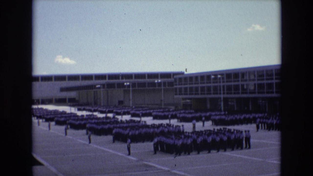 Rows of uniformed students stand across a paved yard beside a long school building.