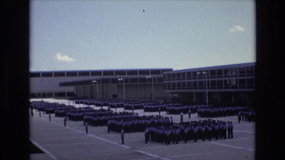 1973 Colorado Springs Super 8 frame showing grouped students standing in formation across a paved schoolyard, with a low modern school building and open sky behind them.