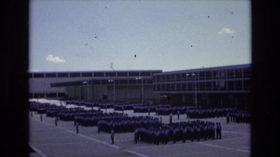 1973 Colorado Springs frame showing large student formations on a paved yard, with a few separated figures standing near the rows and a modern school building in the background.