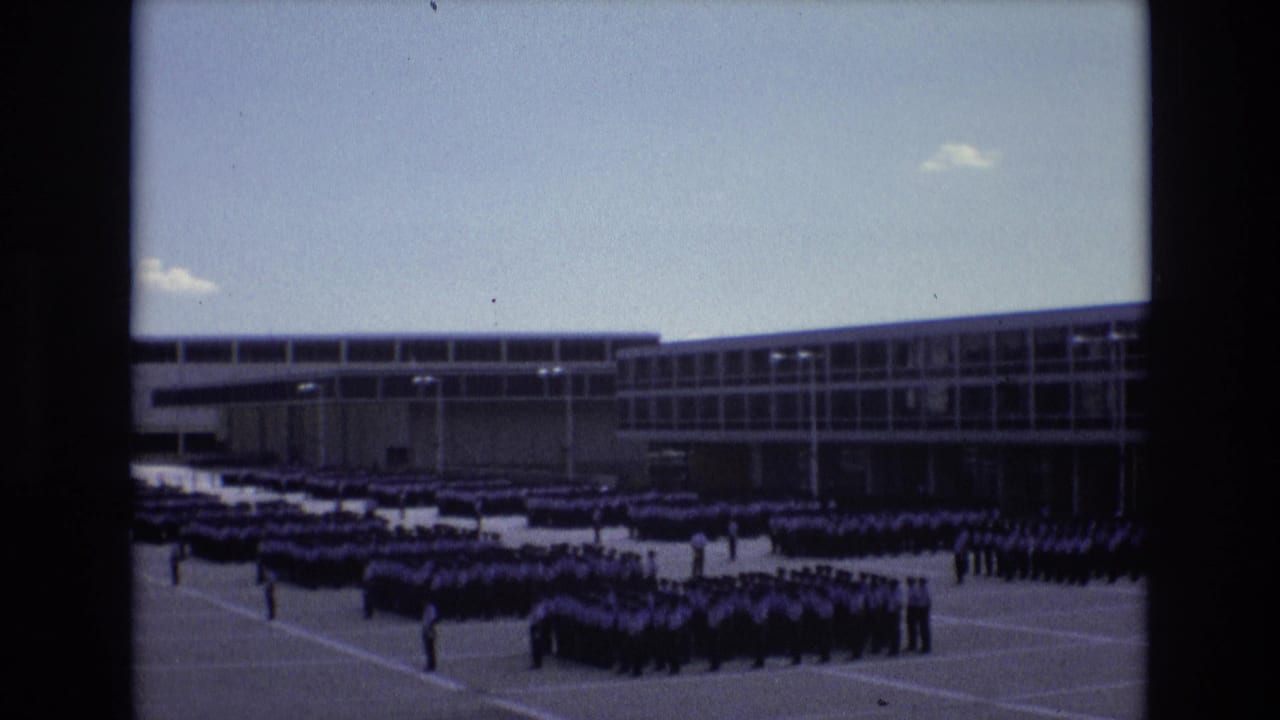 1973 Colorado Springs home-movie frame showing a large distant assembly of uniformed men in ordered rows outside a modern institutional building.