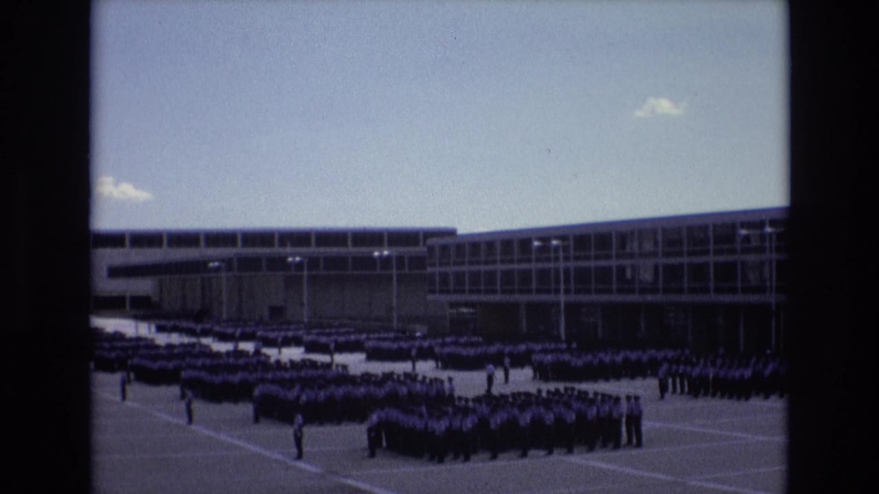 Large formation of uniformed men stands across a courtyard in front of a broad windowed building.