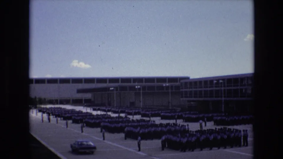 1973 Colorado Springs frame with a classic car crossing the lower foreground while uniformed men remain assembled outside a modern institutional building.