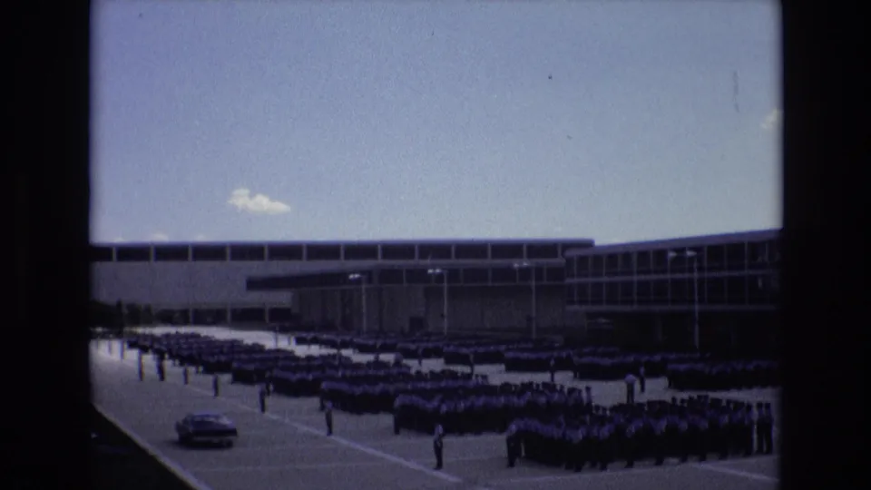Closing 1973 Colorado Springs frame showing a car in the paved foreground and a large uniformed assembly lined up before a glass-fronted institutional building.