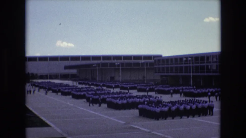 The 1973 Colorado Springs home-movie view holds on cadet formations grouped across a paved courtyard, with the institutional building stretching behind them.