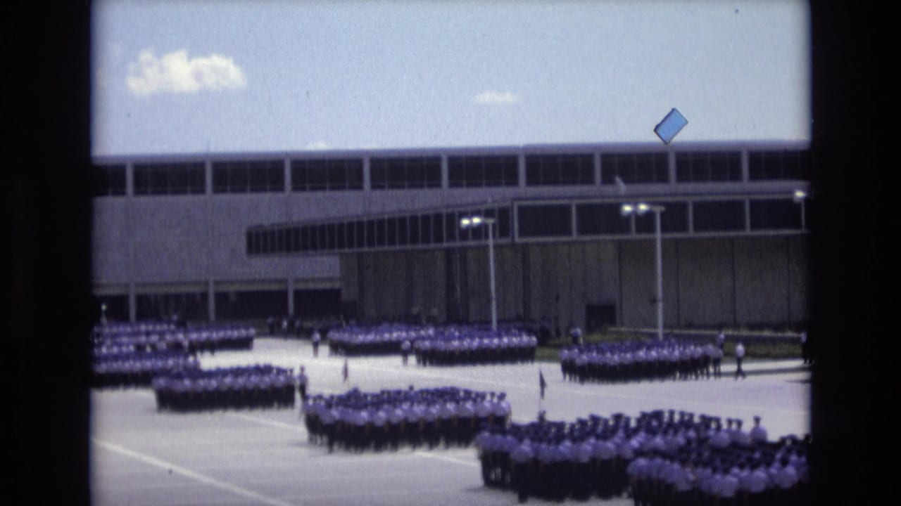 1973 Colorado Springs home-movie frame showing distant uniformed groups arranged in rows on a broad paved area before a large modernist building.