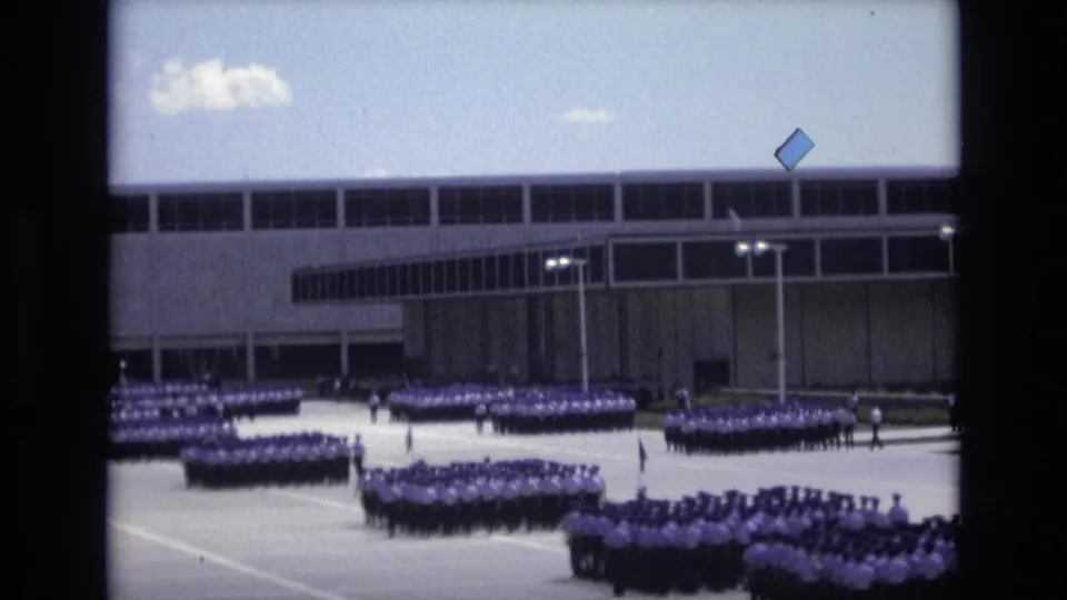 1973 Colorado Springs home-movie frame showing distant uniformed groups arranged in rows on a broad paved area before a large modernist building.