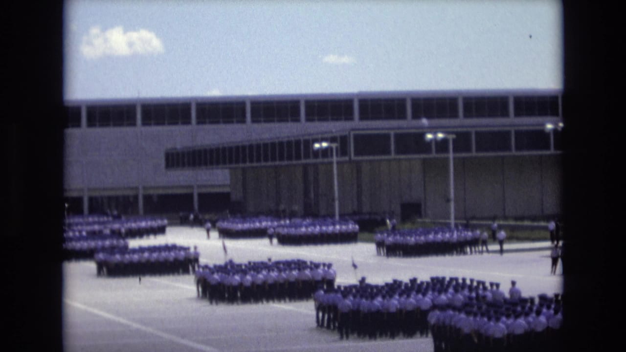Rows of uniformed people gather across a paved parade area in front of a long-windowed building.
