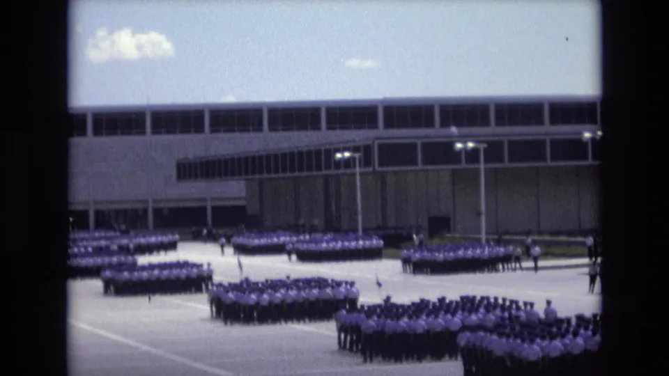 1973 Colorado Springs frame with multiple uniformed formations spread across a paved parade or airfield area near a large building with long bands of windows.