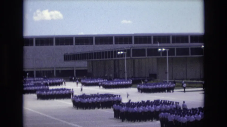 Distant 1973 Colorado Springs 8mm frame showing ordered uniformed groups on a bright paved grounds with a modern institutional building in the background.