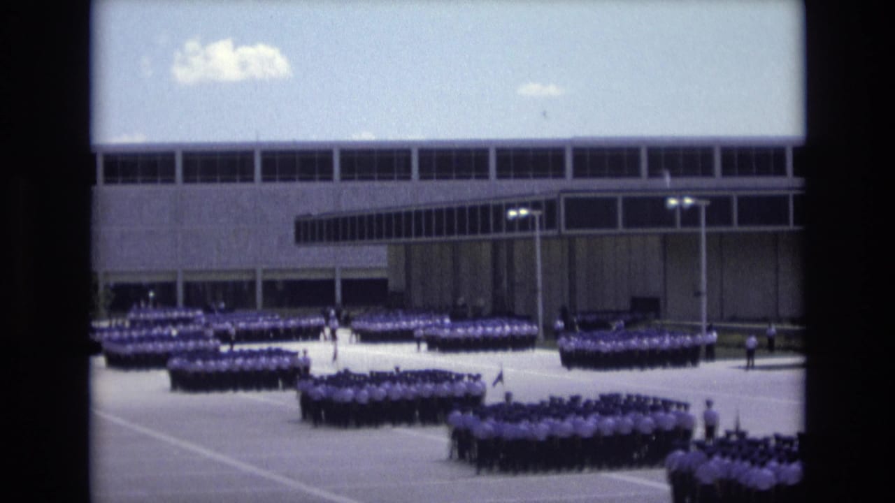 Multiple blocks of uniformed people remain in formation near a modern building.