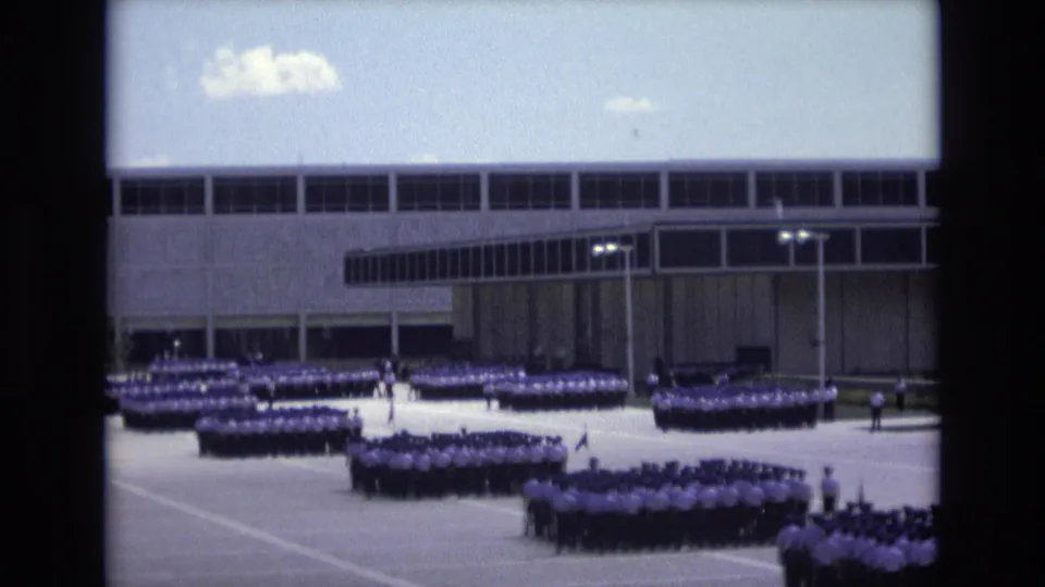 1973 Colorado Springs home-movie still with several uniformed groups standing in ordered blocks on a broad paved area near a large institutional facade.