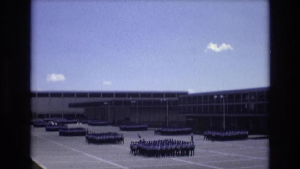 Wider 1973 Colorado Springs home-movie frame showing uniformed groups on a paved courtyard with parked vehicles and a modern institutional building in the background.