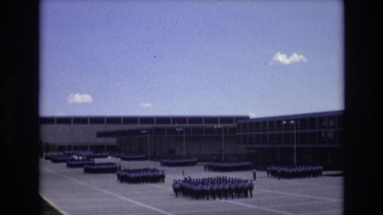 In supplied 1973 Colorado Springs footage, distant uniformed personnel form several rectangular ranks across a large paved plaza in front of a long modern institutional building under a blue sky.