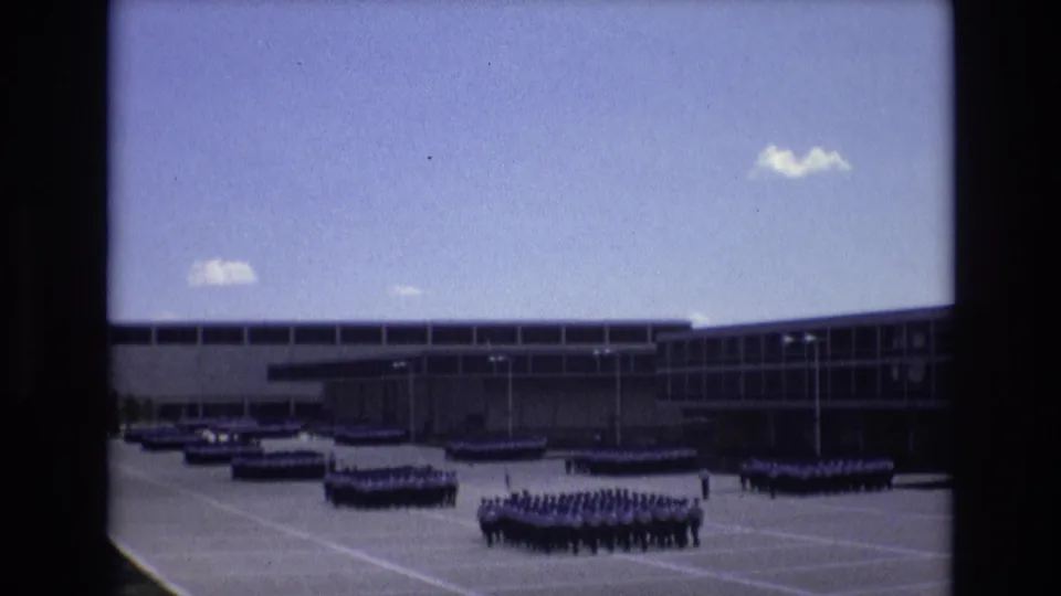 In supplied 1973 Colorado Springs footage, distant uniformed personnel form several rectangular ranks across a large paved plaza in front of a long modern institutional building under a blue sky.