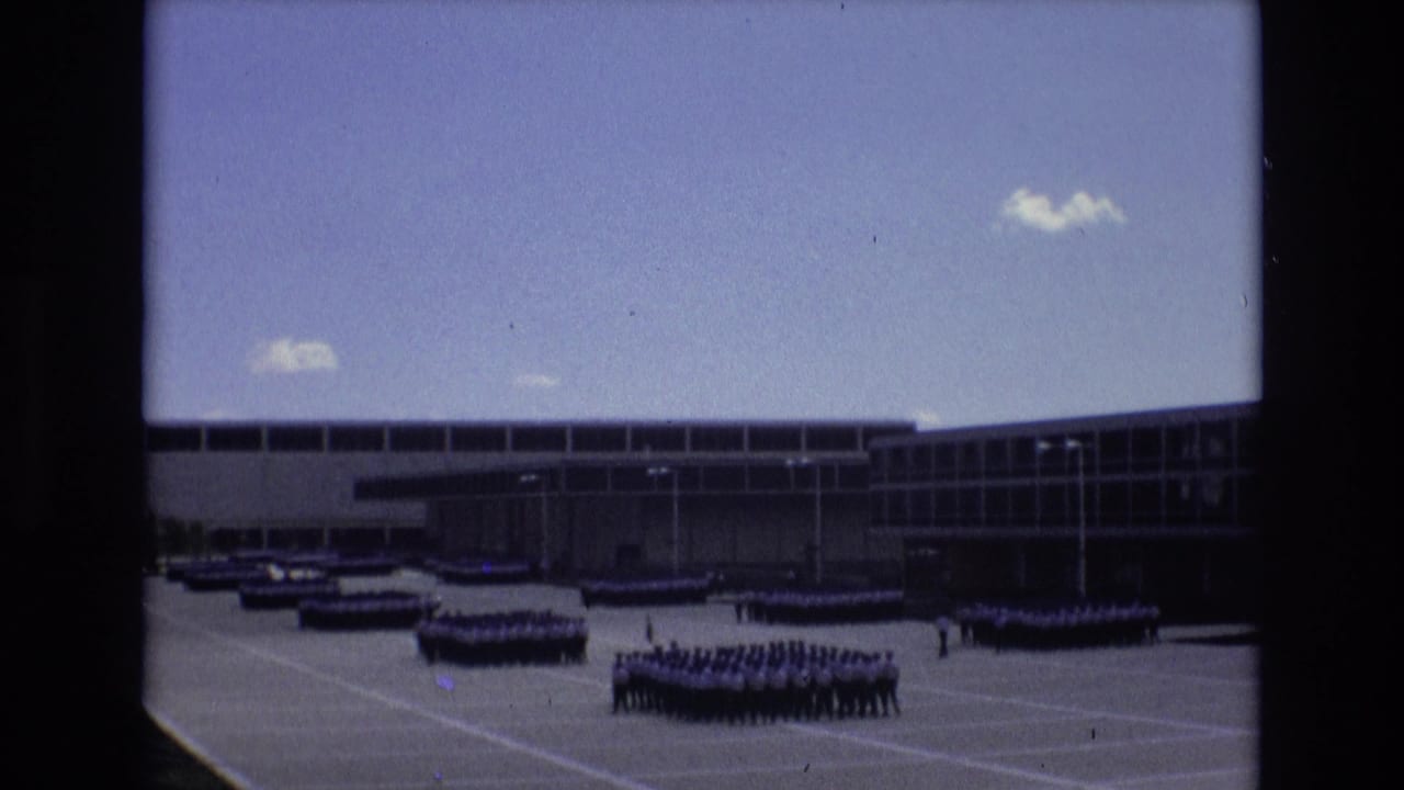 Several uniformed formations are arranged across a paved ground in front of a long building.