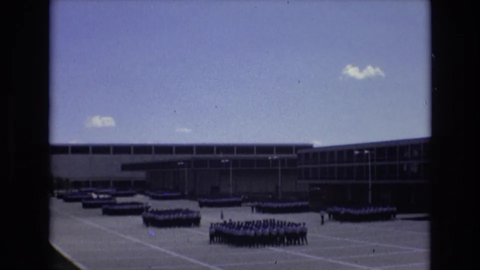 Distant ranks of uniformed personnel appear across the paved parade ground, with the modern building and open sky framing the coordinated maneuver scene in Colorado Springs metadata.